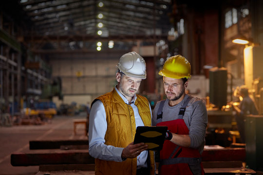 Male foreman and engineer using digital tablet in dark factory