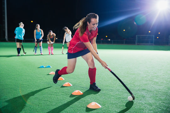 Determined young female field hockey player practicing sports drill on field at night