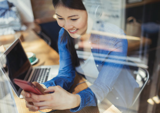 Smiling young woman listening to music headphones texting cell phone at laptop in cafe window