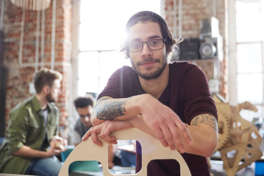 Portrait confident male designer tattoos leaning on prototype in workshop