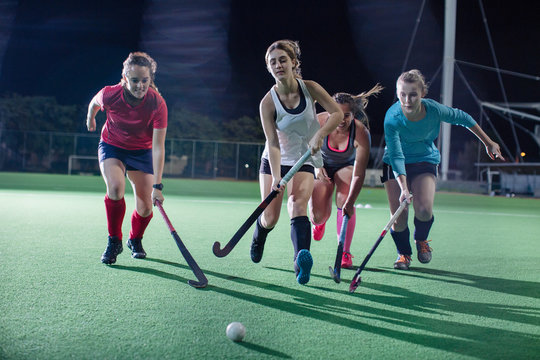 Young female field hockey players running for the ball, playing on field at night