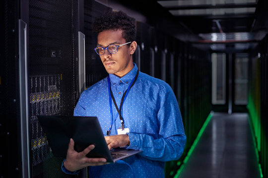 Focused male IT technician using laptop in dark server room