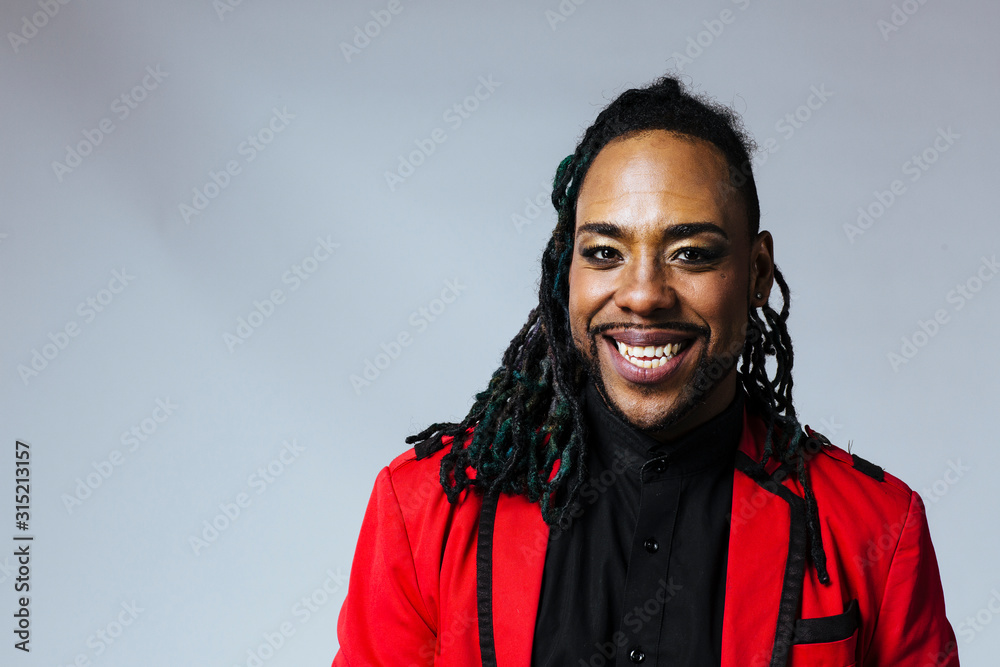 Poster closeup studio portrait of a man wearing a red jacket suit and dreadlocks smiling.