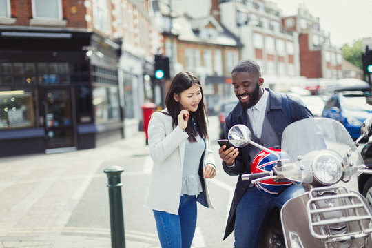 Young couple using cell phone at motor scooter on sunny urban street