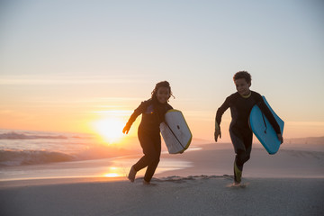 Brother sister in wet suits running boogie boards on summer sunset beach