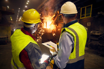 Steelworkers with digital tablet and clipboard talking in steel mill