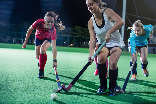 Young female field hockey players running to ball, playing field hockey on field at night