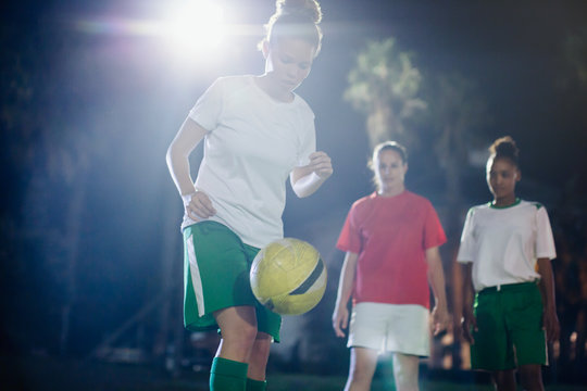 Focused young female soccer player practicing on field at night, kneeing the ball