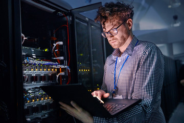 Focused male IT technician working at laptop in dark server room