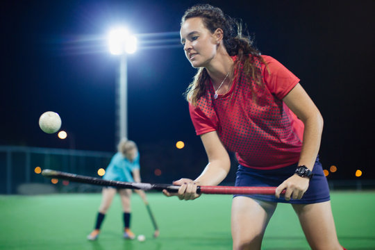 Young female field hockey player bouncing ball off hockey stick, practicing on field at night
