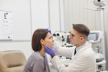 Side view portrait of young ophthalmologist opening eye of female patient while checking her vision in med clinic, copy space