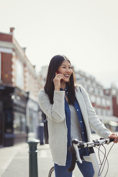 Smiling young woman commuting bicycle, talking on cell phone on sunny urban street