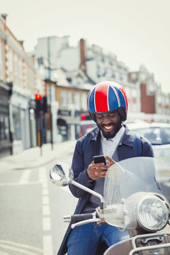 Smiling young businessman in helmet on motor scooter, texting cell phone on urban street