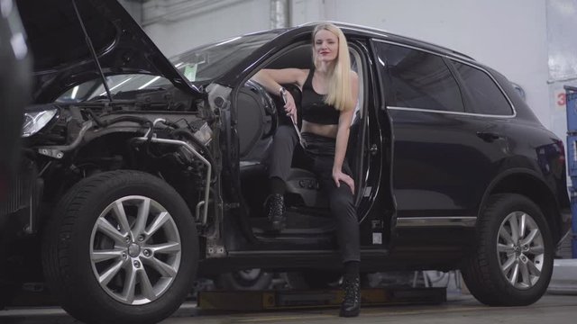 Portrait of attractive Caucasian blond woman sitting at driver's seat with adjustable wrench and smiling. Beautiful auto mechanic posing in broken automobile at workplace.