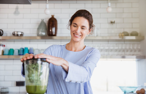 Smiling woman making healthy green smoothie in blender in kitchen
