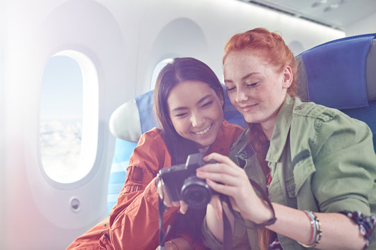 Smiling young women friends looking at photos on digital camera on airplane