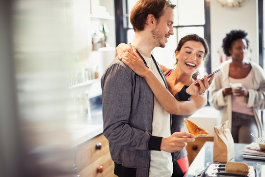 Affectionate couple hugging, texting with smart phone in kitchen
