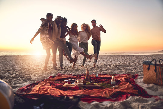Playful young friends kicking, enjoying picnic on sunny summer sunset beach
