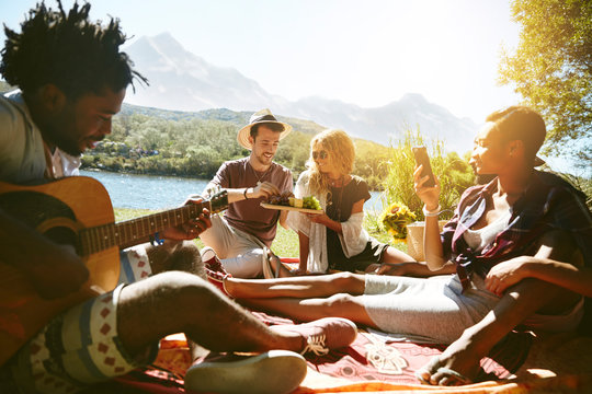 Young friends hanging out, playing guitar enjoying picnic at sunny summer riverside