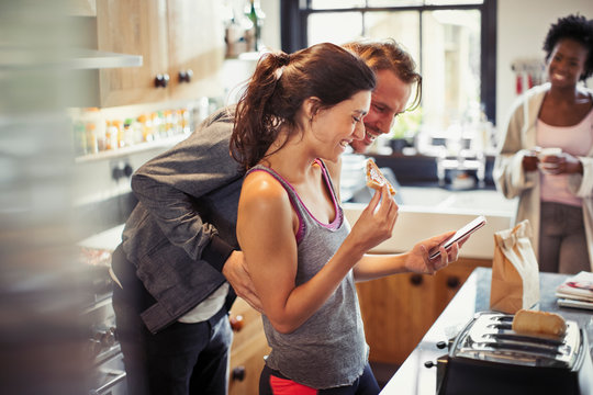 Smiling couple texting with smart phone, eating toast in kitchen - Powered by Adobe