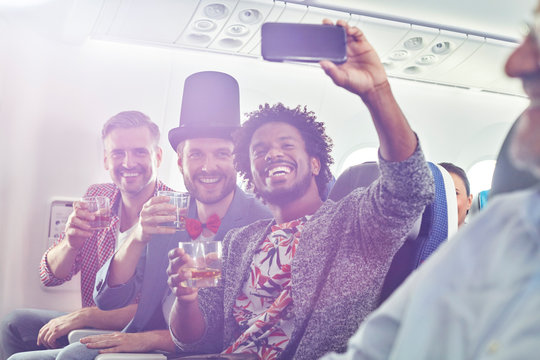 Enthusiastic young male friends camera phone drinking taking selfie on airplane