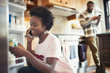 Woman talking on smart phone, reading label on jar in refrigerator in kitchen