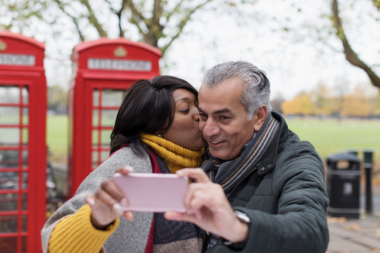 Senior couple kissing and taking selfie in park in front of red telephone booths