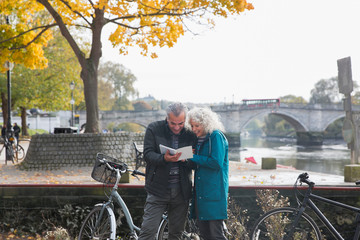 Senior couple with bicycles traveling, looking at guidebook along autumn river