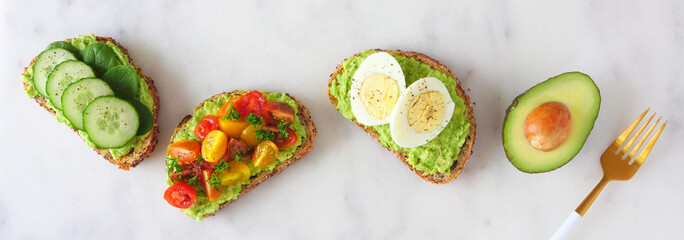 Healthy avocado toasts with eggs, tomatoes and cucumber spinach. Banner orientation, flat lay over a white marble background.