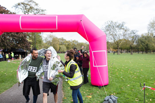 Volunteer wrapping male runners in thermal blanket at marathon finish line