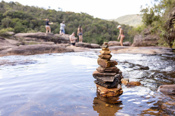 Close up of small rocks stacked on top of each other in a pond part of a stream leading to a waterfall with people out of focus on the edge in the background