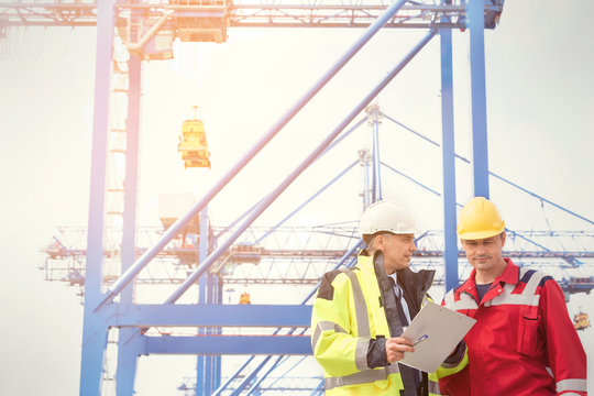 Dock worker and manager with clipboard talking below crane at shipyard