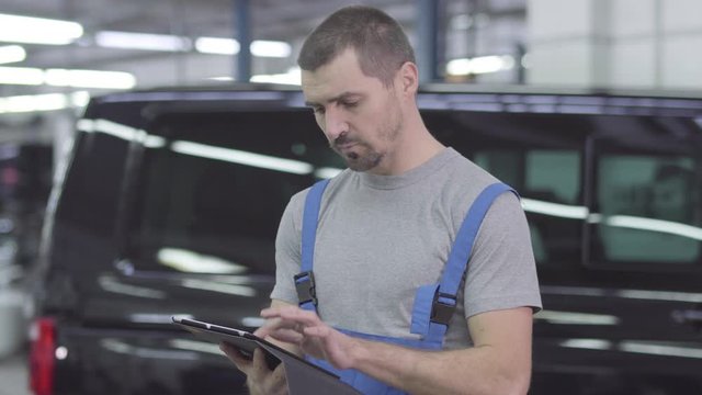 Portrait of young handsome Caucasian man in workwear using tablet, looking at camera and smiling. Confident cheerful male auto mechanic at the background of car. Inspiration, lifestyle, occupation.