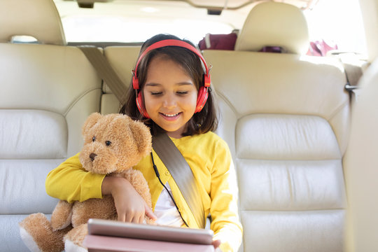 Smiling girl teddy bear using digital tablet headphones in back seat of car