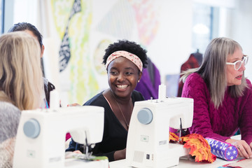 Happy female fashion designer working, talking colleague at sewing machine