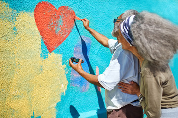 Senior couple painting heart-shape mural on sunny wall