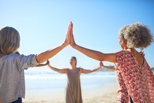 Women joining hands in circle on sunny beach during yoga retreat