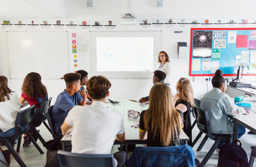 High school students watching female teacher leading lesson at projection screen in classroom