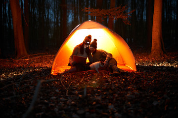 Couple using digital tablet inside glowing tent in woods at night