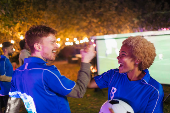 Happy friends cheering, watching soccer match on projection screen in backyard