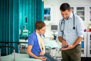 Male pediatrician showing digital tablet to boy patient with arm in sling in hospital