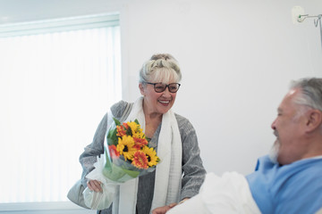 Happy senior woman bringing flower bouquet to husband recovering in hospital room