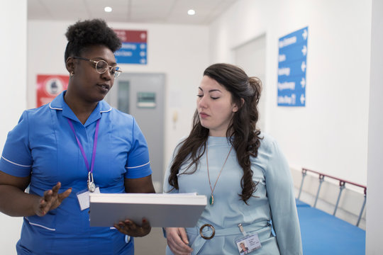 Female doctor and nurse making rounds, discussing medical chart in hospital corridor