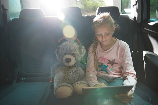 Girl with teddy bear using digital tablet in back seat of car