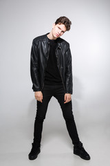 Caucasian young guy posing in studio, full-length portrait of a model in black clothes. Handsome teenager in a black jacket, jeans and sneakers on a gray background. Man in a leather jacket