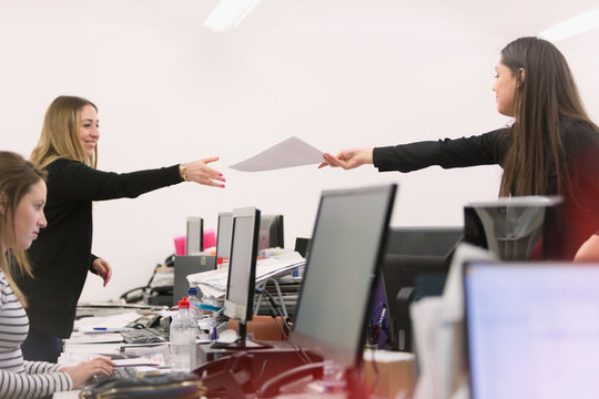 Businesswoman handing paperwork to colleague over computers in office
