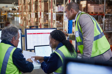 Managers meeting reviewing paperwork at laptop and computer in distribution warehouse