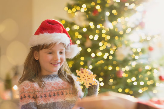 Girl in Santa hat holding snowflake ornament in front of Christmas tree