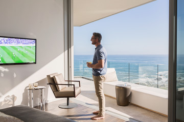 Man watching soccer on TV at sunny luxury patio doorway with ocean view