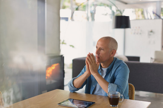 Pensive man with digital tablet drinking coffee and looking away near wood burning stove fireplace